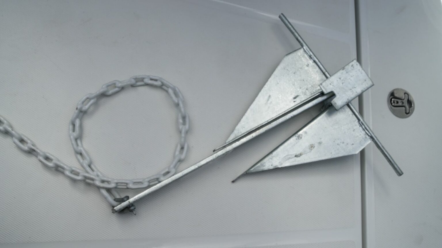 License-free image - A metal boat anchor with a fluke design lies on a white surface next to a coiled chain. The anchor has a long shank, two flat triangular flukes, and a stock across the top. The chain is made of metal links joined together. A fluke, danforth, or lightweight anchor.