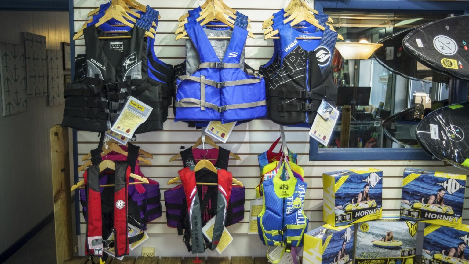 License-free image A display of various life jackets is hung on a wall inside a store. The life jackets come in different sizes and colors, including blue, black, red, and yellow. There are also a few water sports boards displayed on the right side.