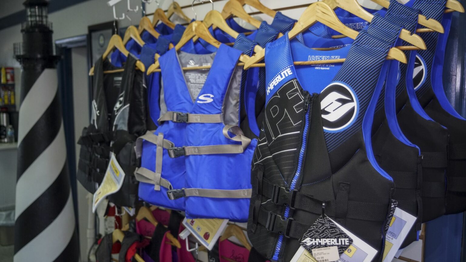 License-free image - Several life jackets in blue and black colors hang on display racks in a store. The jackets have brand names and logos visible, with tags attached. A spiral-striped lighthouse decoration stands in the background on the left side.