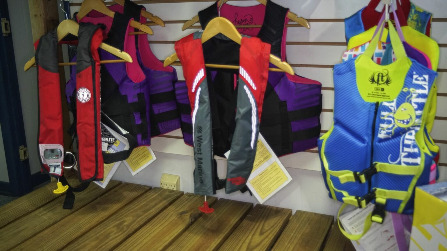 License-free image - A variety of life jackets are hanging on wooden hangers against a slatted wall display. The life jackets come in different colors, including red, purple, grey, and blue, and appear to be for both adults and children. Informational tags are attached to them.