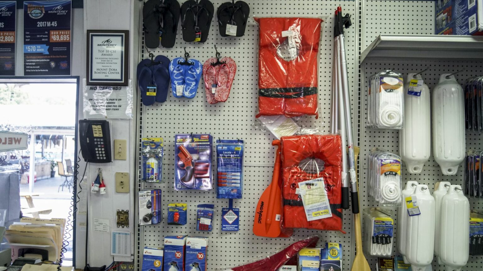 License-free image -  A store display featuring various boating supplies including life jackets, paddles, ropes, flip-flops, and other accessories. Items are neatly arranged on a pegboard and shelves. A wall-mounted telephone and framed certificates are seen on the left side.