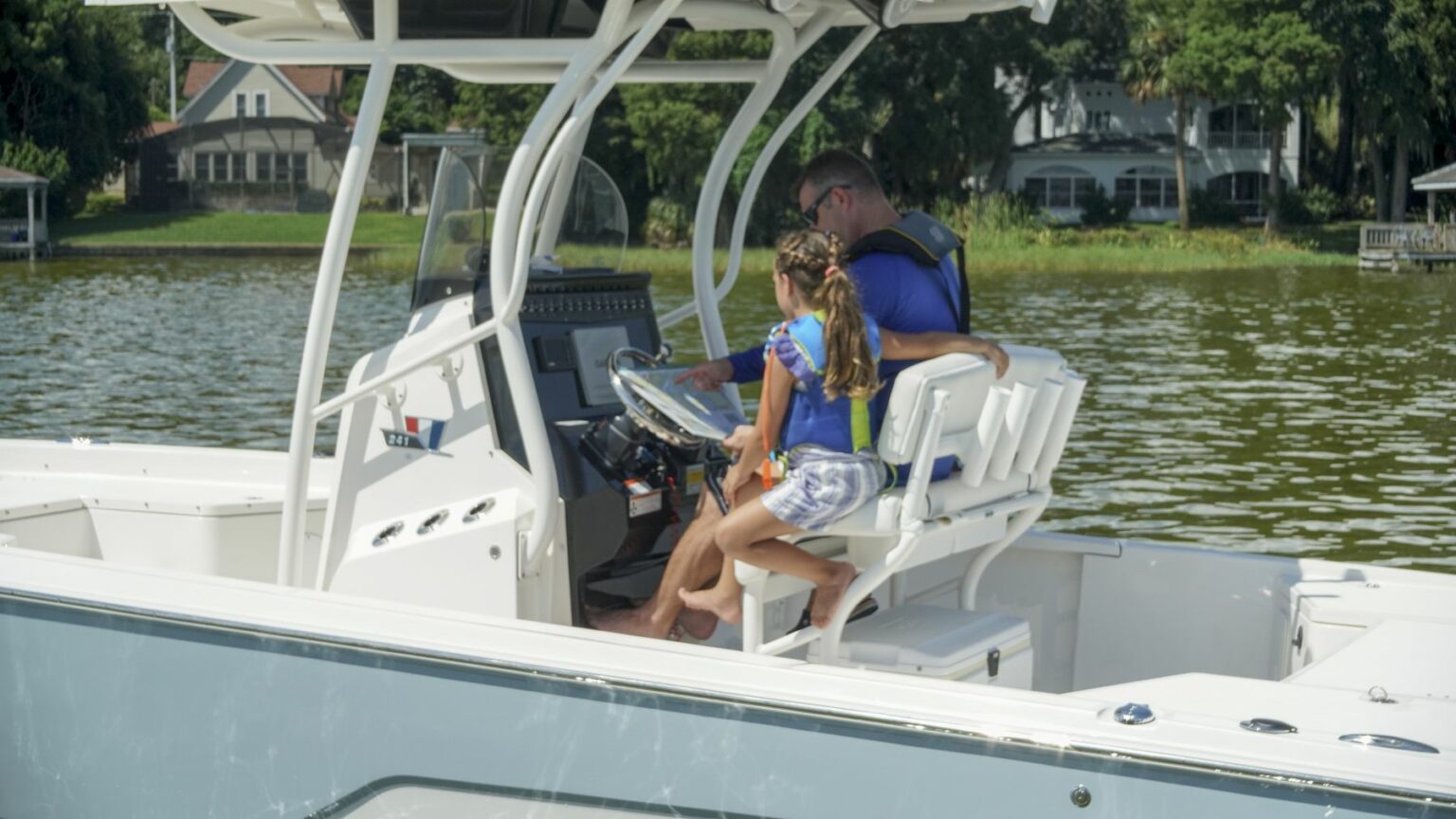 License-free image - A man and a young girl are sitting on a white boat in a lake. The man is steering while the girl, wearing a swimsuit and a life jacket, looks at the controls. The background shows houses and trees on the lake shore. Using paper nautical charts aboard a small boat.