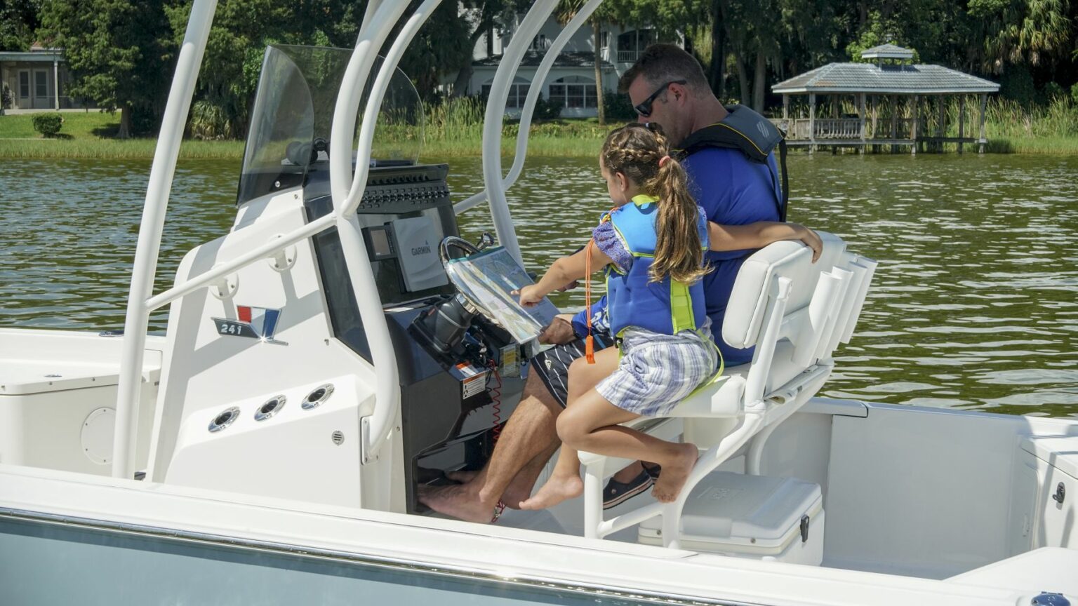 License-free image - A man and a young girl wearing life jackets are sitting together on a boat. The man is steering while the girl looks on, with her hands on the controls. They are on a calm lake, and trees and a gazebo are visible in the background. Using paper nautical charts aboard a small boat.