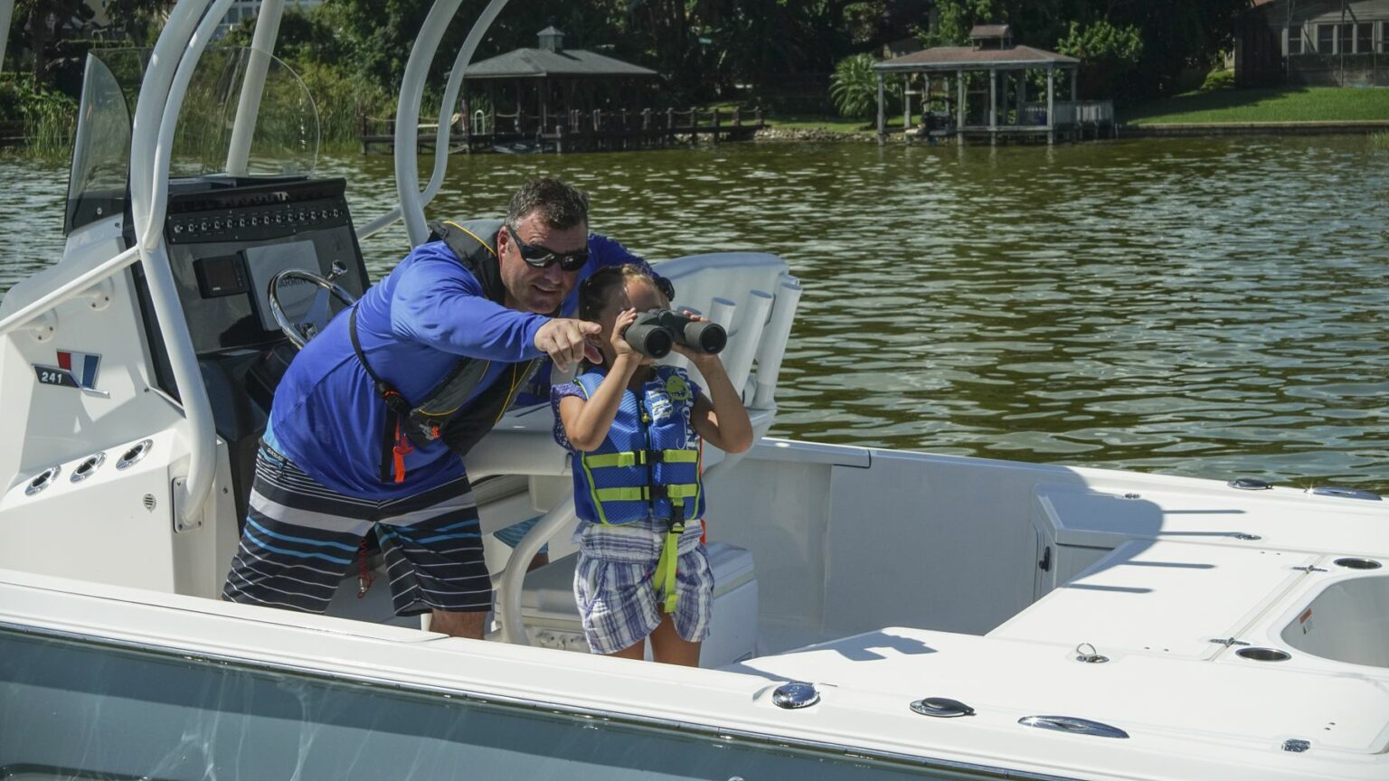 License-free image - A man wearing a blue long-sleeve shirt and sunglasses is pointing out something in the distance to a child using binoculars. They are both wearing life jackets and standing on a white boat with a calm body of water and shoreline in the background. A boater making a weather observation while boating.