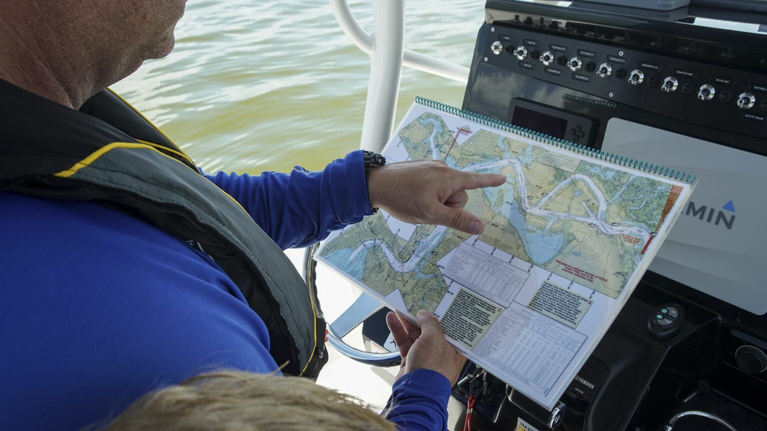 Royalty-free image - Two people, wearing life jackets, are on a boat. One person is pointing at a navigational chart displayed on a laptop or device near the boat&#039;s control panel, which shows a body of water with various channels and landmarks. Using paper nautical charts aboard a small boat.