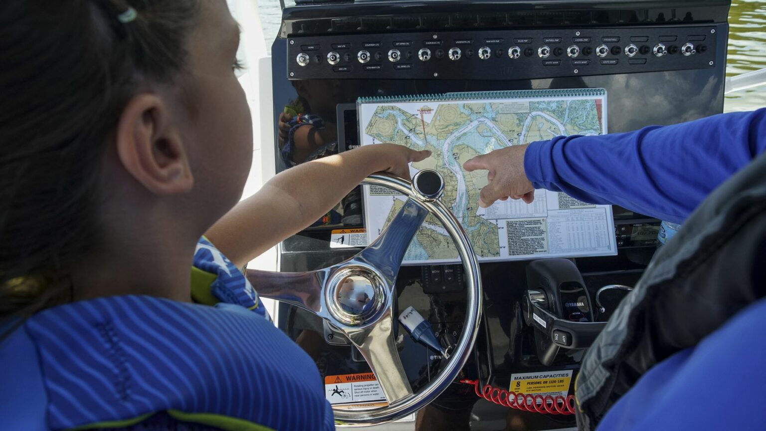 License-free image - A person points to a map on a boat&#039;s dashboard while another person steers. Both are wearing blue clothing, and one has a life vest on. The map shows a detailed layout of waterways. The boat&#039;s control panel and steering wheel are in view. Using paper nautical charts aboard a small boat.