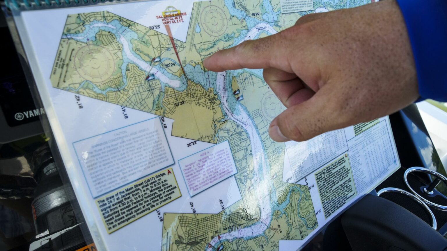 License-free image - A hand points to a specific location on a navigational map. The map contains various waterways, symbols, and text boxes for nautical information. The corner of a boat&#039;s instrument panel is visible in the background. Using paper nautical charts aboard a small boat.