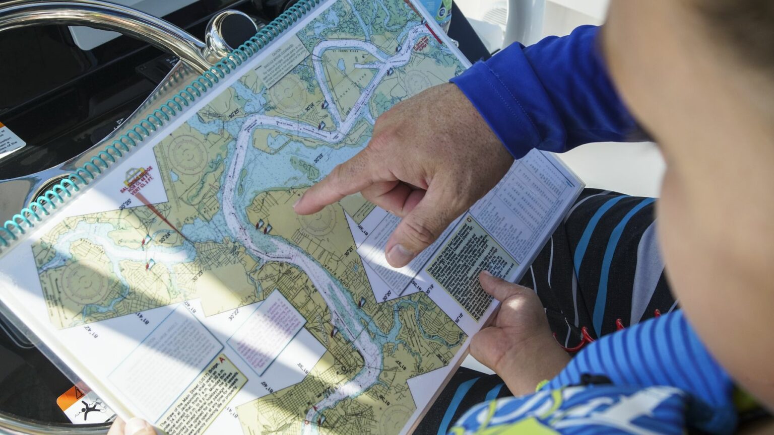 License-free image - A person points to a location on a detailed navigational waterway map in a spiral-bound book. The map shows rivers, channels, and surrounding land. The person, wearing a blue shirt, is likely planning or discussing a route. Another individual looks on. Using paper nautical charts aboard a small boat.