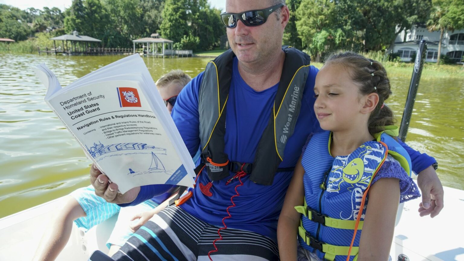 Royalty-free image A man wearing sunglasses and a life jacket sits on a boat, holding a U.S. Coast Guard manual. A girl in a life jacket sits beside him, looking at the book. Another person, partially hidden, is seen behind the man. The boat is on a lake with trees in the background.