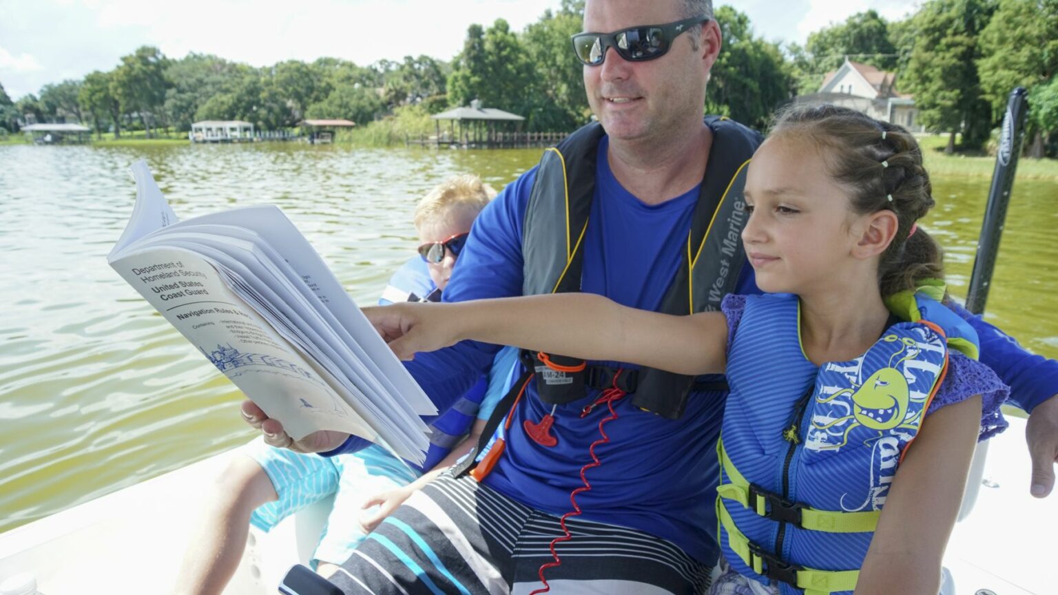 License-free image A man and a young girl wearing life vests are sitting on a boat on a lake. The girl is pointing at a page in a book that they are holding together. Another child is partially visible in the background. The lake is surrounded by trees.