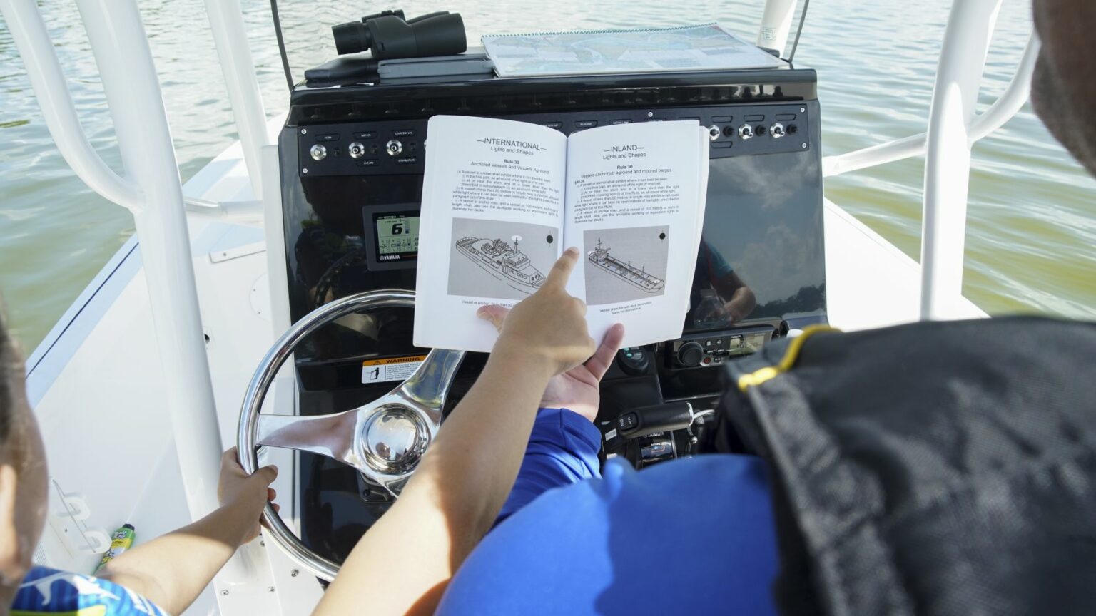 License-free image A person reviewing the United States Coast Guard Navigation Rules and Regulations Handbook open, pointing at diagrams while another person, possibly a child, holds the boat&#039;s steering wheel. The vessel&#039;s console with instruments and water in the background are visible, indicating that they are on a boat.