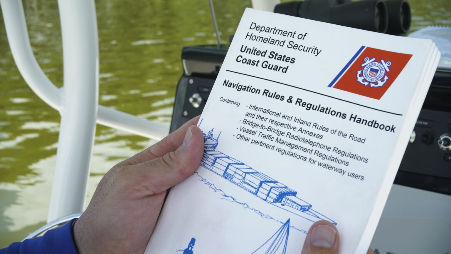 Royalty-free image A person holds a book titled &quot;Department of Homeland Security, United States Coast Guard: Navigation Rules &amp; Regulations Handbook.&quot; The book contains information on navigation safety, rules of the road, and other regulations. The background shows a boat and water.