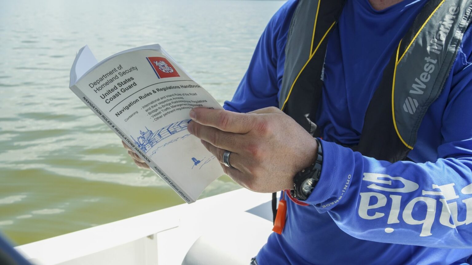 License-free image A person wearing a blue long-sleeve shirt and a life jacket reads a booklet titled &quot;Department of Homeland Security United States Coast Guard: Navigation Rules &amp; Regulations Handbook&quot; while sitting in a boat on the water.