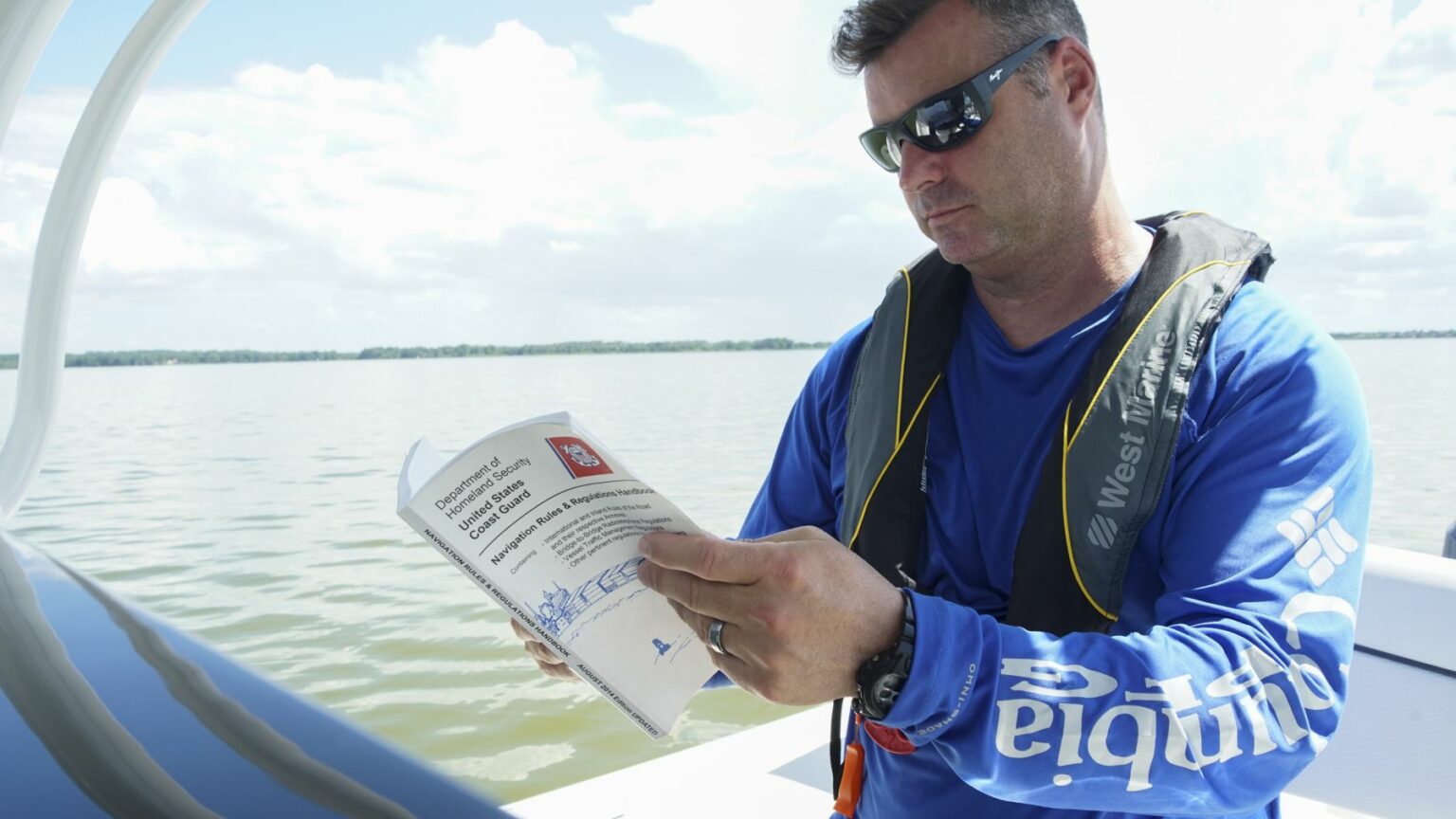 License-free image A man wearing sunglasses and a life jacket studies a chart while on a boat, with a calm body of water and a cloudy sky visible in the background. He is wearing a blue long-sleeve shirt and holding a navigation manual.