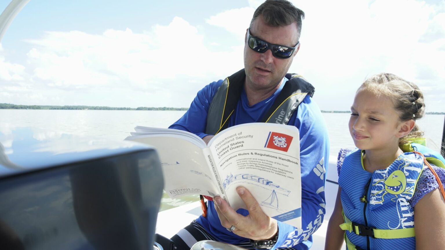 License-free image A man wearing sunglasses and a life jacket reads a boating safety manual while sitting on a boat. Next to him, a young girl wearing a blue life jacket with marine motifs appears to be listening attentively. They are both on a calm body of water under a sunny sky.