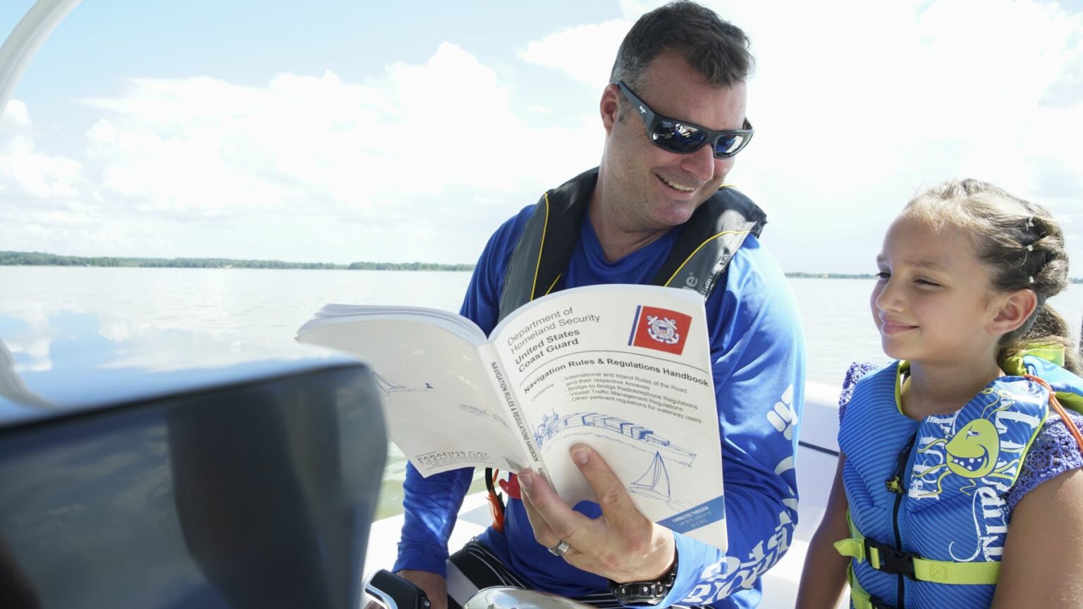 License-free image A man wearing sunglasses and a life jacket reads a boating safety manual while sitting on a boat. Next to him, a young girl wearing a blue life jacket with marine motifs appears to be listening attentively. They are both on a calm body of water under a sunny sky.