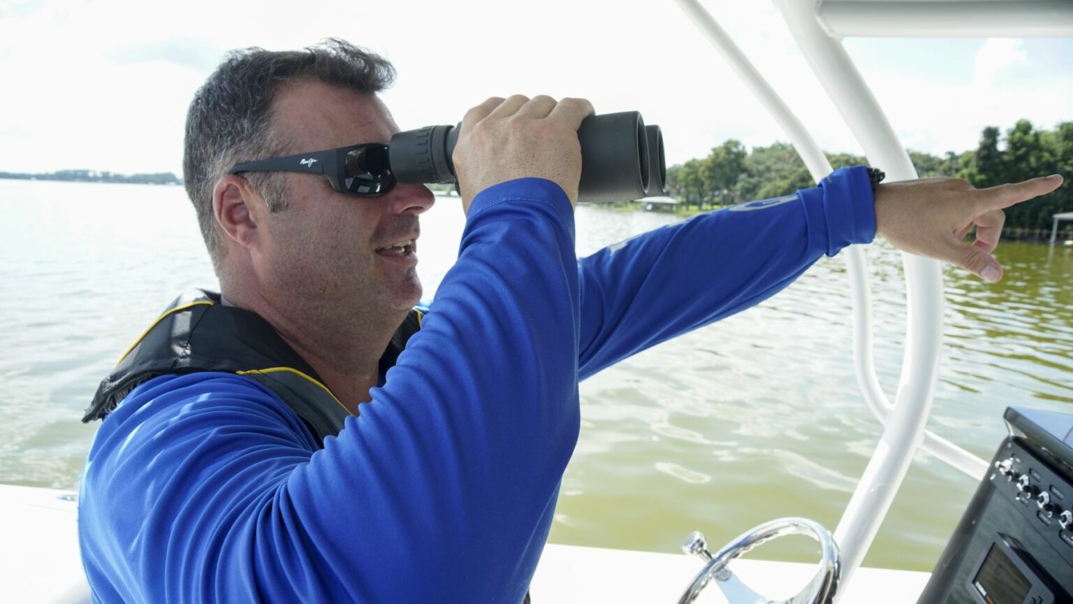 License-free image - A man on a boat is looking through binoculars. He is wearing a blue long-sleeve shirt, sunglasses, and a life vest. With his left hand, he is pointing towards something in the distance over the water. The background shows a calm body of water and some greenery. A boater making a weather observation while boating.