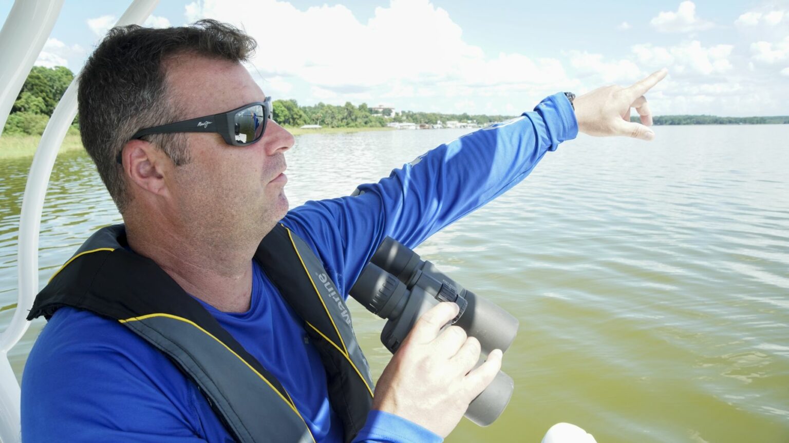 License-free image - A man wearing sunglasses and a blue shirt holds binoculars in one hand and points out across a calm body of water with his other hand. He is standing on a boat, and the background includes a clear sky with scattered clouds and a distant shoreline. A boater making a weather observation while boating.