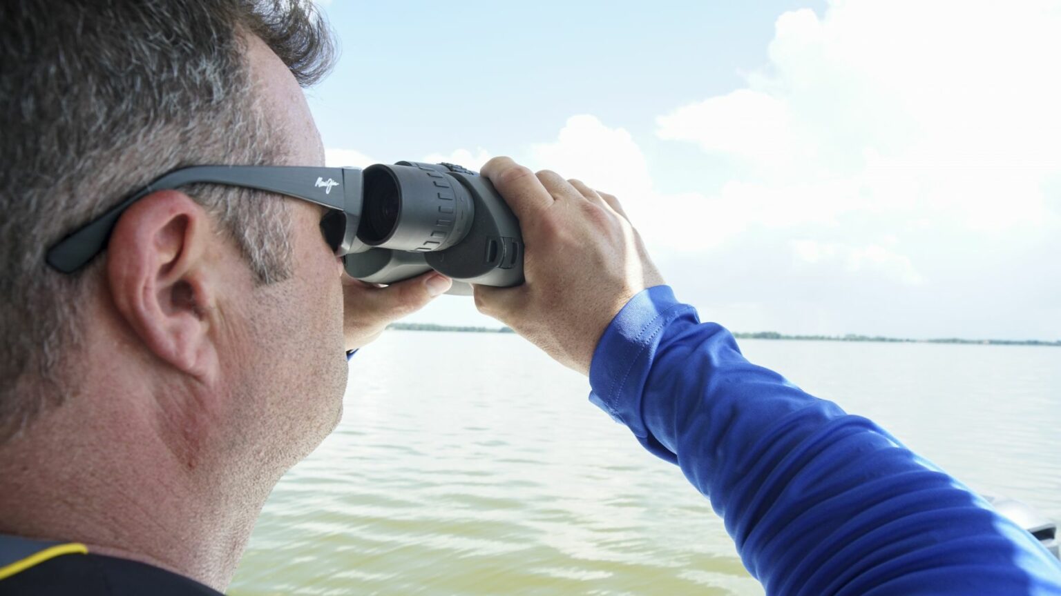 Royalty-free image - A person in a blue long-sleeve shirt and sunglasses is looking through binoculars over a calm body of water with a relatively clear sky, suggesting a scene of boating or bird-watching. The focus is on the person&#039;s back and binoculars. A boater making a weather observation while boating.