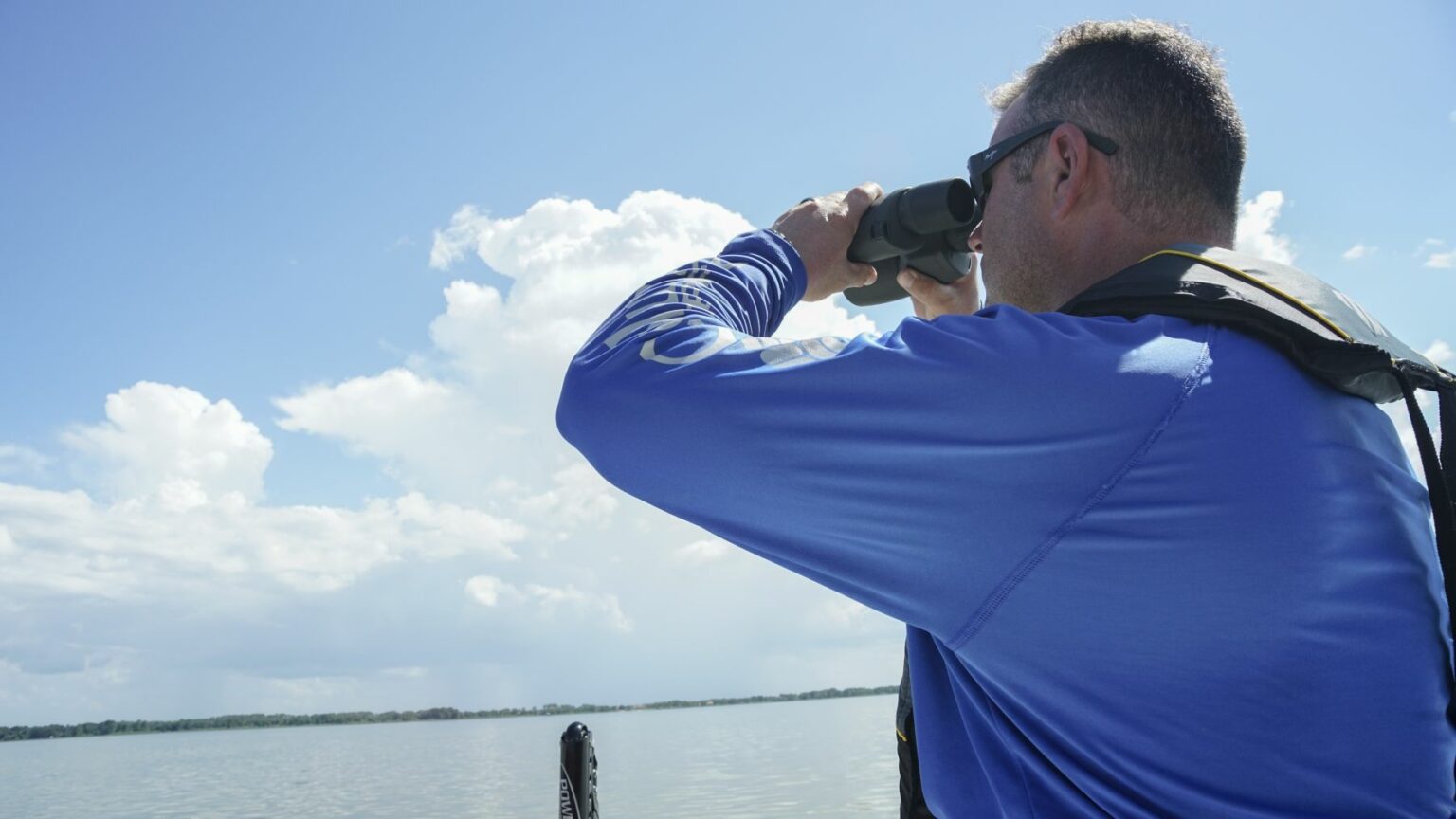 License-free image - A person wearing a blue shirt and life jacket holds binoculars while looking out over a large body of water. The sky is mostly clear with a few clouds. The scene appears to be peaceful and serene.