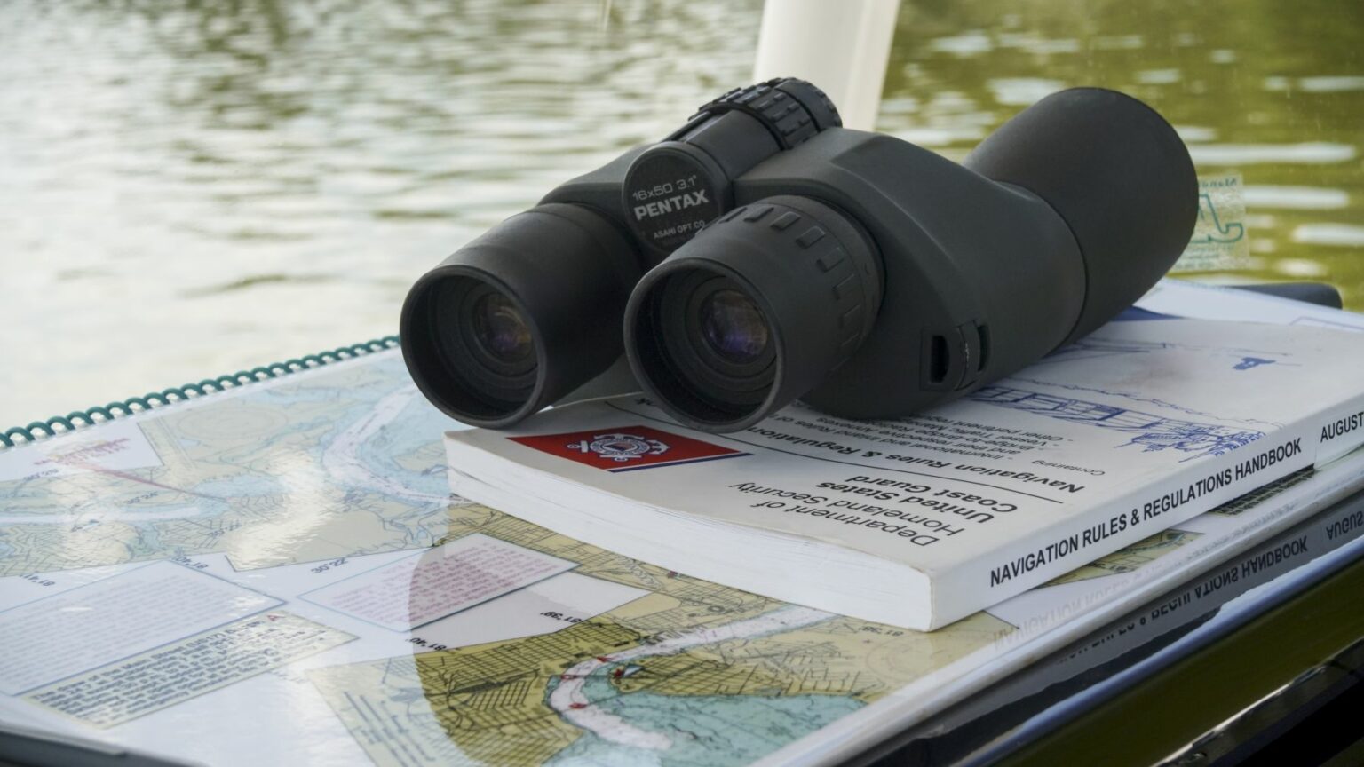 License-free image - A pair of binoculars rests on top of a navigational chart and a handbook titled &quot;Navigation Rules &amp; Regulations Handbook.&quot; The background includes a view of the water, indicating a maritime setting. Binoculars for making a weather observation while boating.