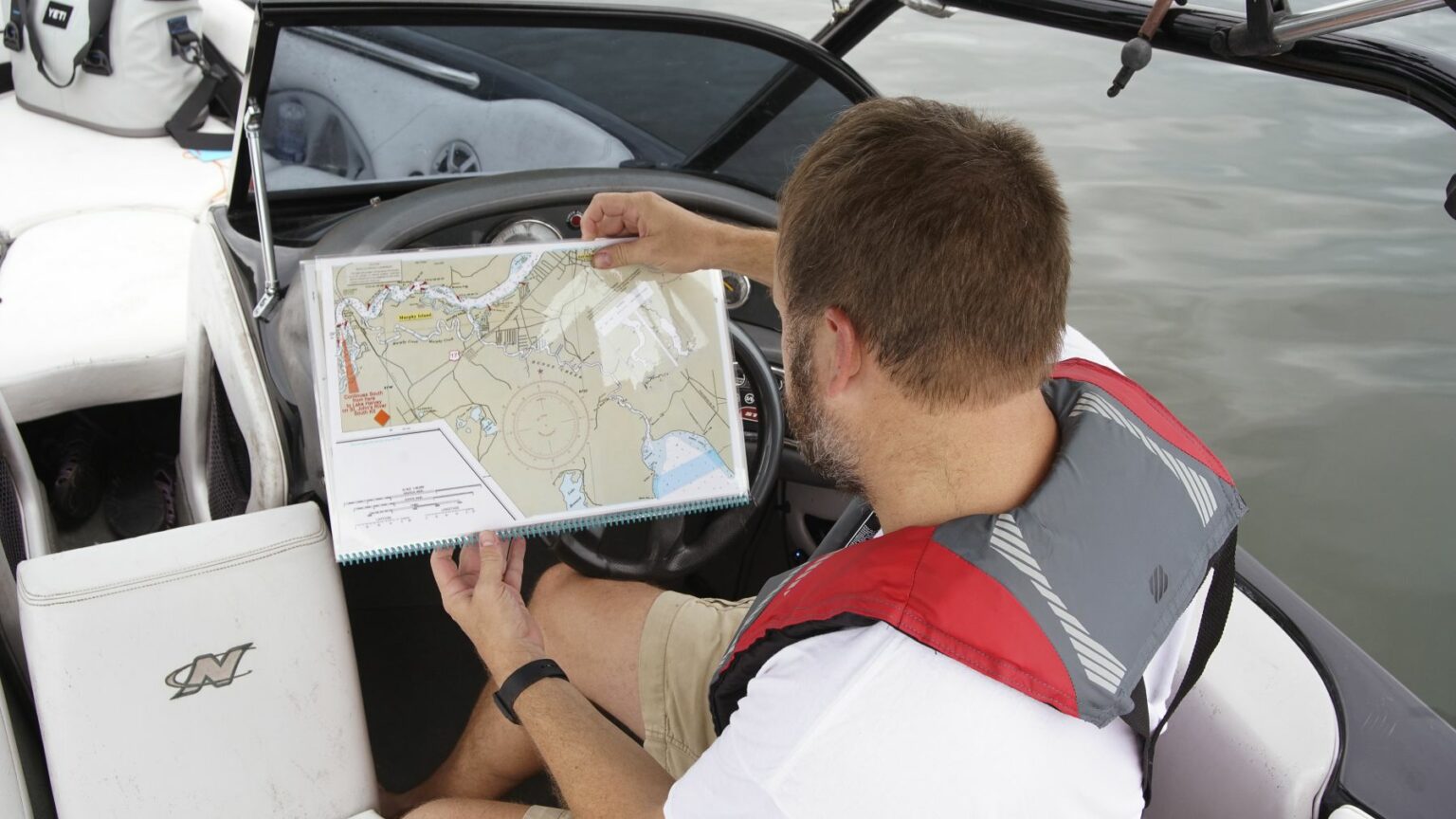 Royalty-free image - A man wearing a life jacket sits in a boat, holding and examining a nautical map. The water is visible in the background, and the boat&#039;s steering wheel is nearby.