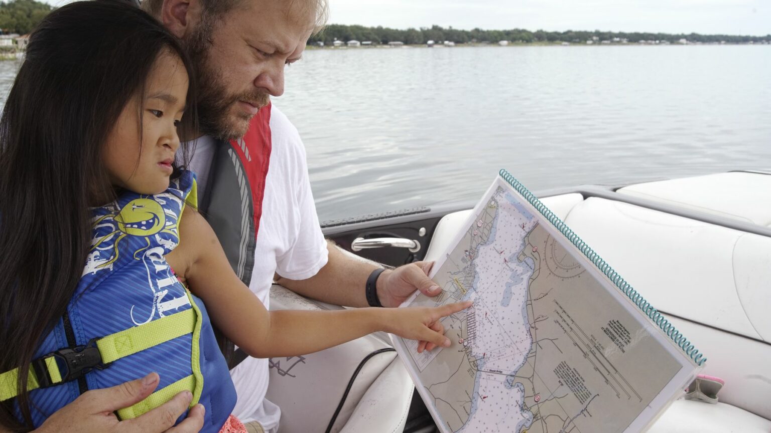 Royalty-free image - A man and a child are sitting in a boat, looking at a large map. The child is pointing at a spot on the map. They both wear life jackets, and the water and shoreline are visible in the background.