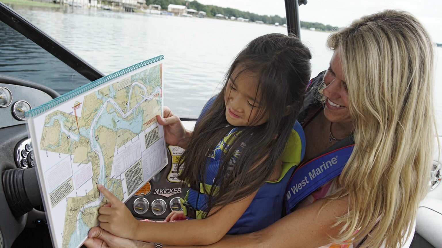 Royalty-free image - A woman and a child are sitting in a boat, looking at a nautical chart together. They are both wearing life jackets. The boat is on a calm body of water with trees and houses in the background.