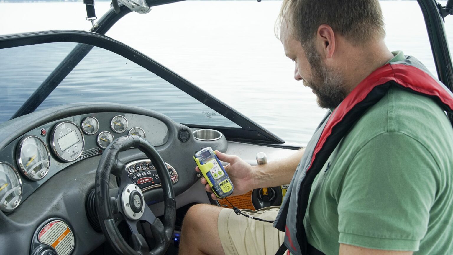 Royalty-free image - A man in a green shirt and life vest is sitting in a boat, intently examining his handheld EPIRB device. The boat&#039;s dashboard displays various gauges and a steering wheel, while the surrounding water remains calm and serene.