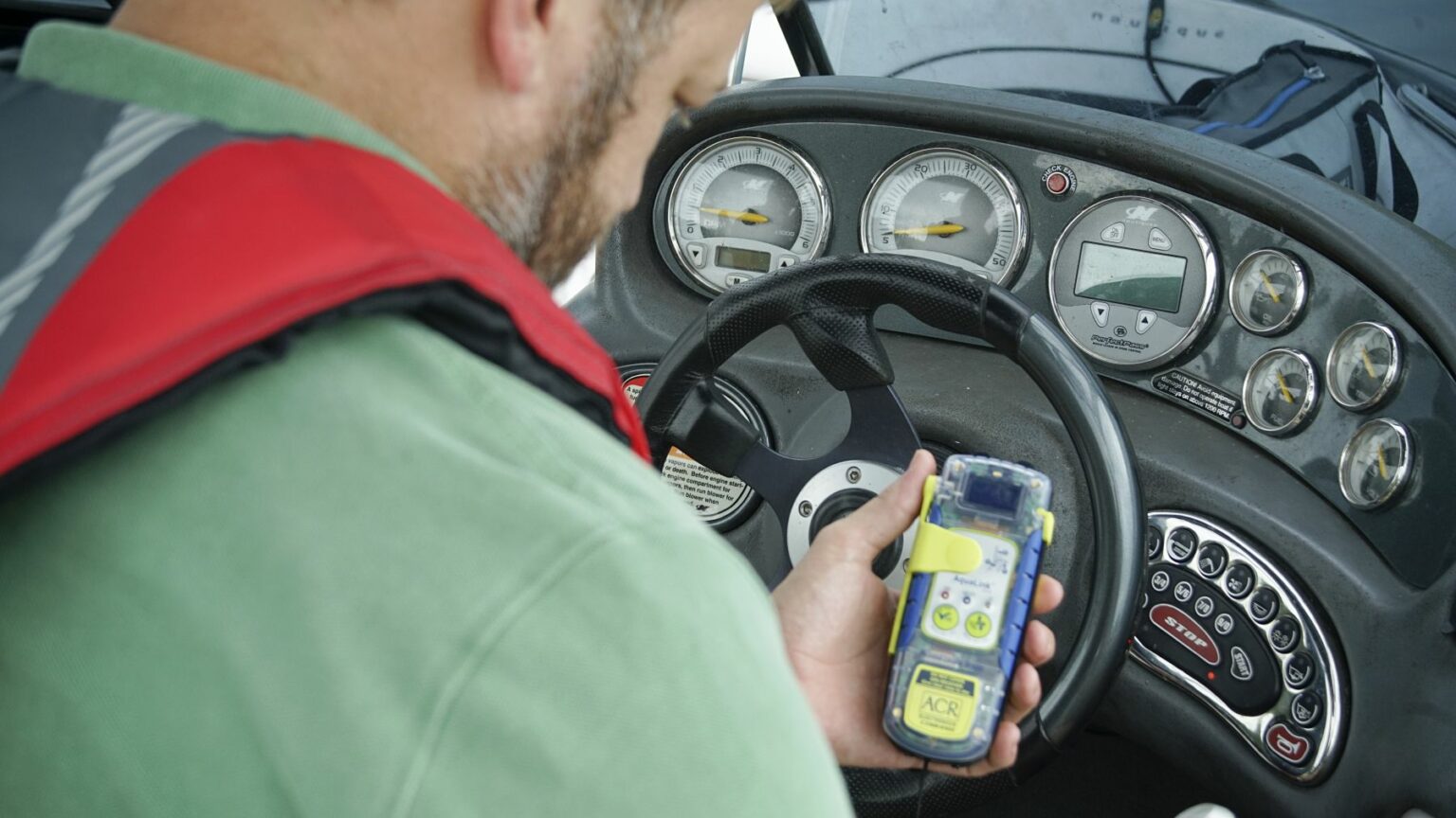 Royalty-free image - A person is holding a handheld electronic device, resembling a personal locator beacon (PLB), with both hands inside a cockpit. The device features a screen and buttons, surrounded by multiple dials and gauges on the panel near the steering wheel.