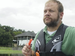 License-free image - A man with a beard wearing a green shirt and a life jacket holds an orange whistle while standing near a body of water. Behind him, trees and a white building with a dark roof are visible under a cloudy sky. Using a whistle to attract attention aboard a boat.