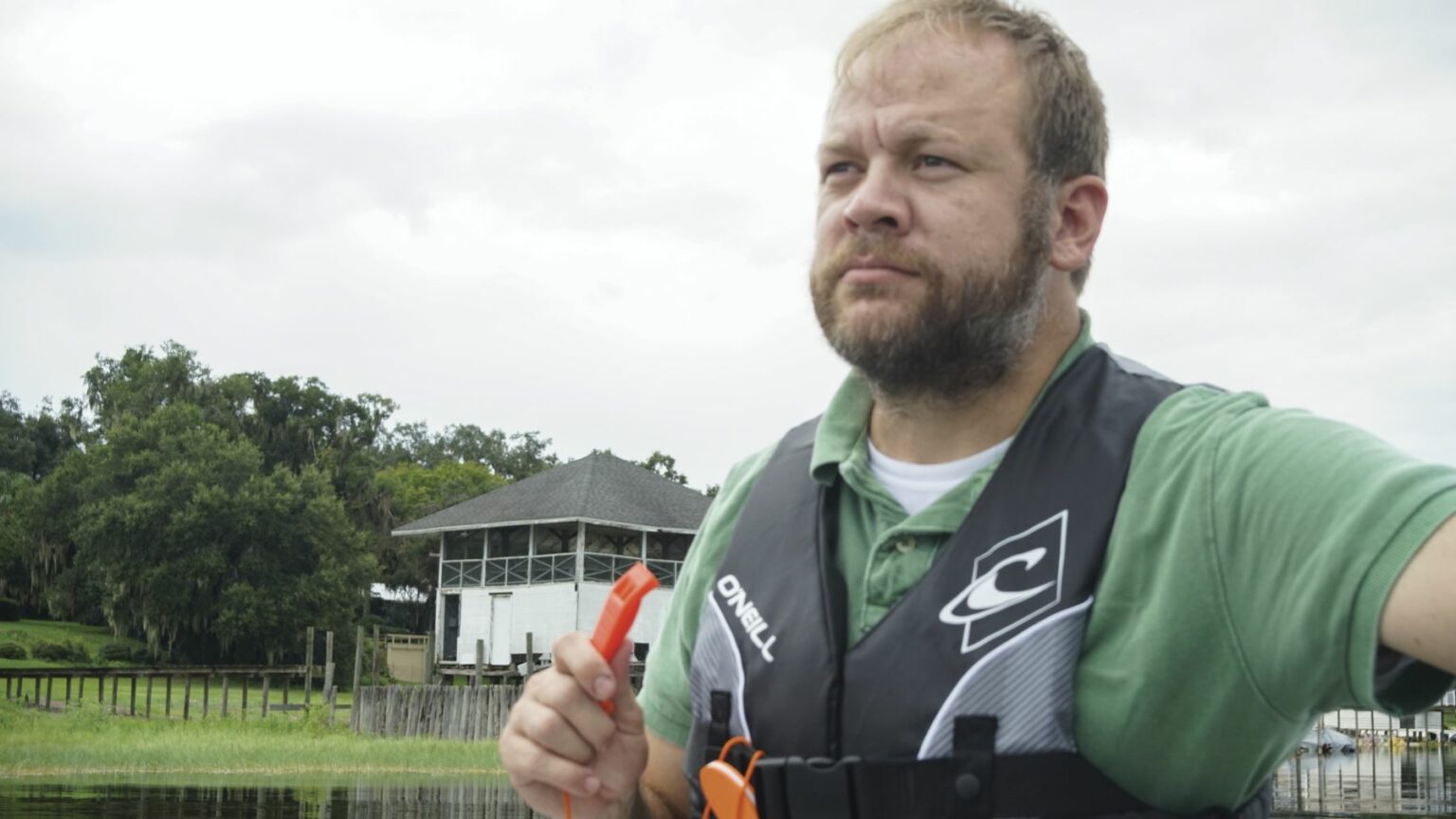 License-free image - A man with a beard wearing a green shirt and a life jacket holds an orange whistle while standing near a body of water. Behind him, trees and a white building with a dark roof are visible under a cloudy sky. Using a whistle to attract attention aboard a boat.