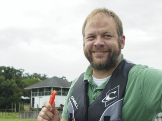 License-free image - A man with short hair and a beard is smiling while standing outdoors. He is wearing a green shirt and a black life jacket, holding an orange whistle. The background features a white building with trees and a cloudy sky. Using a whistle to attract attention aboard a boat.