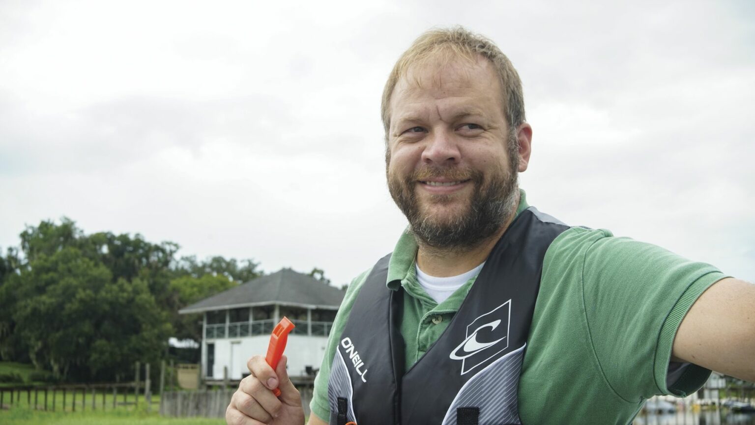 License-free image - A man with short hair and a beard is smiling while standing outdoors. He is wearing a green shirt and a black life jacket, holding an orange whistle. The background features a white building with trees and a cloudy sky. Using a whistle to attract attention aboard a boat.