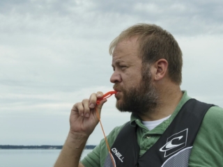 License-free image - A man with short hair and a beard is standing outdoors wearing a green shirt and a life vest. He is blowing a red whistle with a cord attached. The background shows a calm body of water and a cloudy sky. Using a whistle to attract attention aboard a boat.