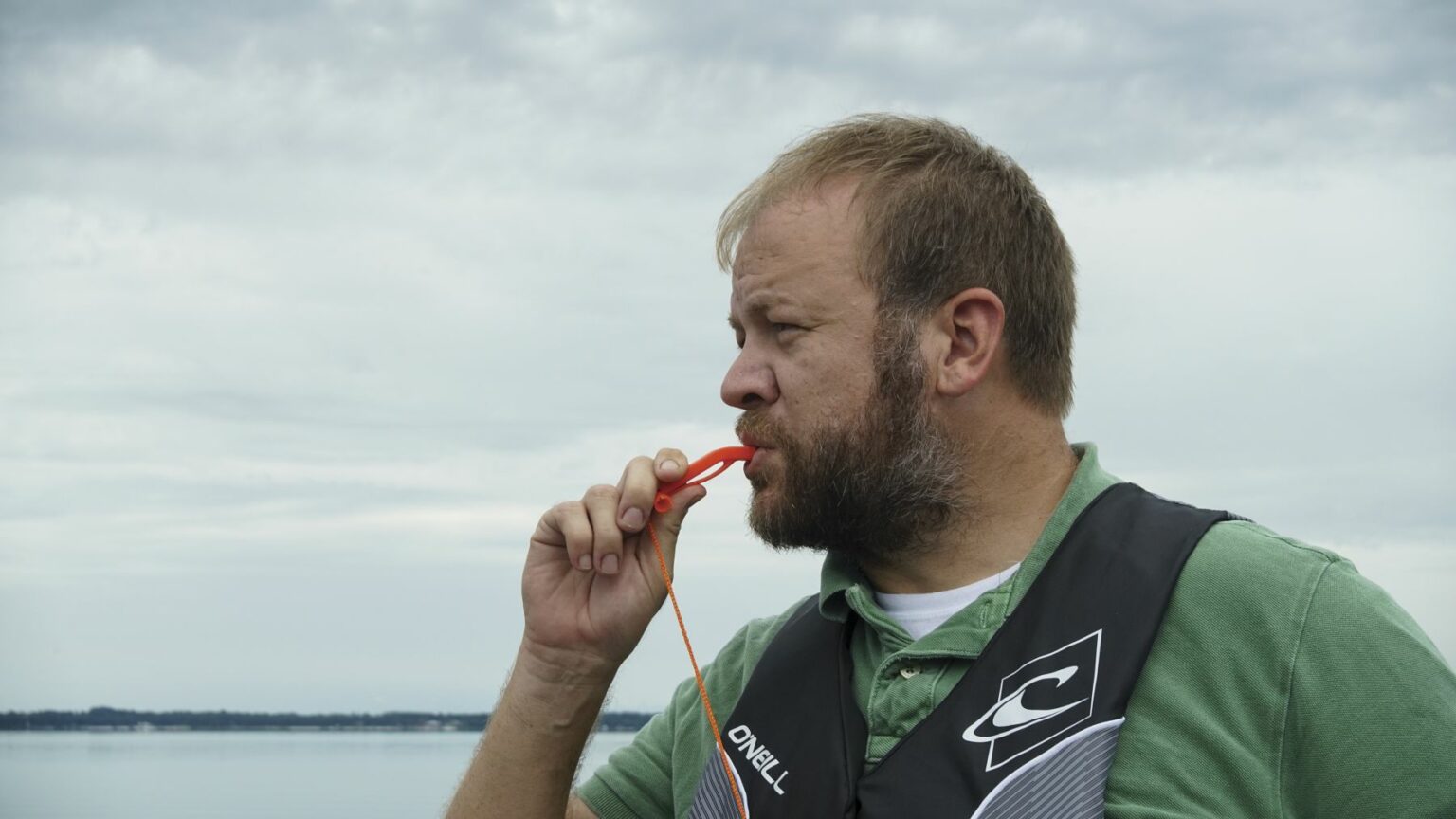 License-free image - A man with short hair and a beard is standing outdoors wearing a green shirt and a life vest. He is blowing a red whistle with a cord attached. The background shows a calm body of water and a cloudy sky. Using a whistle to attract attention aboard a boat.