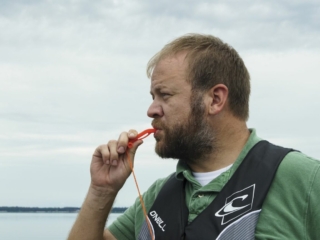 License-free image - A man with a beard and short hair stands in front of a body of water, blowing a whistle. He is wearing a green shirt and a black life jacket. The sky is overcast, and the water appears calm. Using a whistle to attract attention aboard a boat.