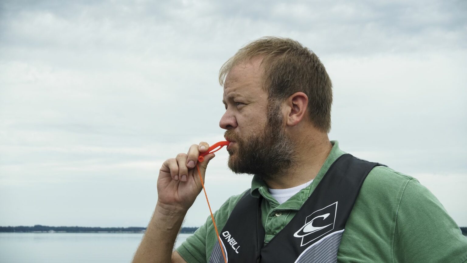 License-free image - A man with a beard and short hair stands in front of a body of water, blowing a whistle. He is wearing a green shirt and a black life jacket. The sky is overcast, and the water appears calm. Using a whistle to attract attention aboard a boat.