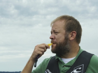 License-free image - A man with a beard is standing outdoors under a cloudy sky, wearing a green shirt and a black and white life jacket. He appears to be blowing a yellow whistle. The background shows a distant, blurred landscape. Using a whistle to attract attention aboard a boat.