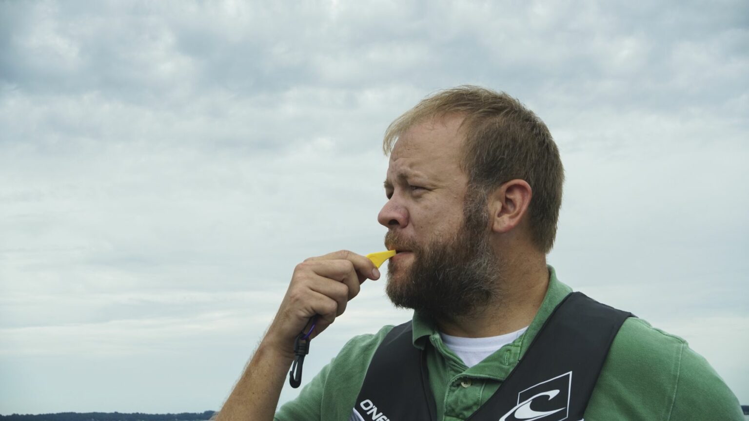 License-free image - A man with a beard is standing outdoors under a cloudy sky, wearing a green shirt and a black and white life jacket. He appears to be blowing a yellow whistle. The background shows a distant, blurred landscape. Using a whistle to attract attention aboard a boat.