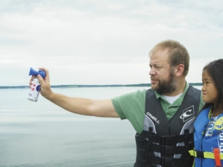 License-free image - A man in a life jacket is holding an air horn, pointing it out towards the water. Beside him stands a young girl in a life jacket, looking in the same direction. The background features a calm lake under an overcast sky. Using a horn to signal while aboard a boat.