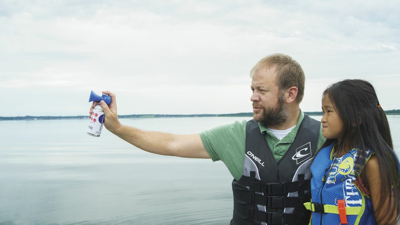 License-free image - A man in a life jacket is holding an air horn, pointing it out towards the water. Beside him stands a young girl in a life jacket, looking in the same direction. The background features a calm lake under an overcast sky. Using a horn to signal while aboard a boat.