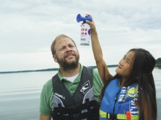 License-free image -  A man with a beard wearing a life jacket stands beside a young girl in a life jacket. The girl is holding up an air horn and appears to be about to activate it. They are both on a calm body of water with a cloudy sky in the background. Using a horn to signal while aboard a boat.