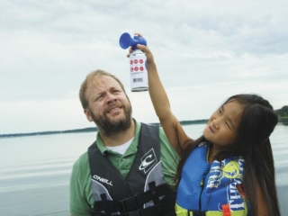 License-free image - An adult in a green shirt and life vest stands on a body of water with a concerned look. A child in a blue life vest holds an air horn above the adult&#039;s head. The background shows a calm lake and an overcast sky. Using a horn to signal while aboard a boat.
