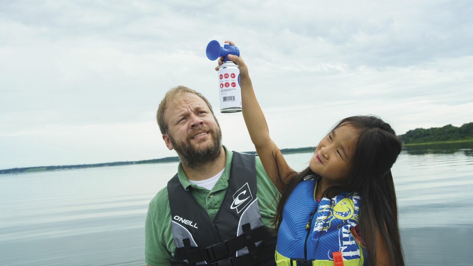 License-free image - An adult in a green shirt and life vest stands on a body of water with a concerned look. A child in a blue life vest holds an air horn above the adult&#039;s head. The background shows a calm lake and an overcast sky. Using a horn to signal while aboard a boat.