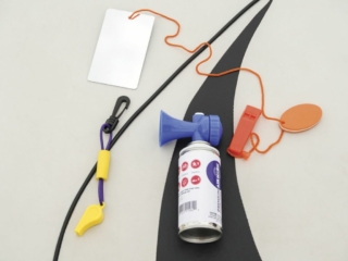 License-free image - Various safety equipment is laid out on a white surface. Items include a blue and white canister with a horn attached, an orange whistle, a white rectangular card with an orange string, and a black cord with a yellow and purple clip. Sound producing devices used to attract attention while boating.