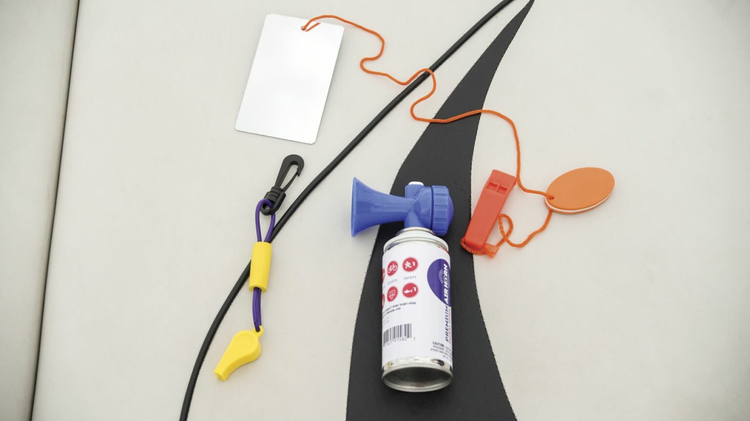 License-free image - Various safety equipment is laid out on a white surface. Items include a blue and white canister with a horn attached, an orange whistle, a white rectangular card with an orange string, and a black cord with a yellow and purple clip. Sound producing devices used to attract attention while boating.