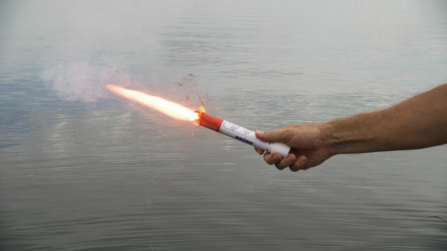 License-free image - A person holds a lit handheld flare emitting bright light and smoke over calm waters, against a backdrop of a slightly cloudy sky. Using a flare during the day.