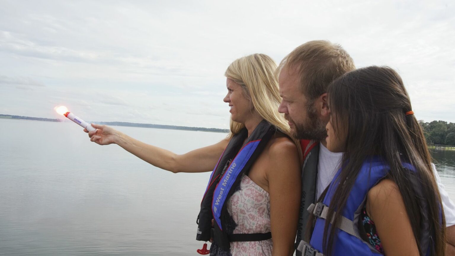 License-free image -  A woman holding a lit flare stands by the water, accompanied by a man and a girl. All three are wearing life jackets and looking toward the flare. The background features a calm body of water and a cloudy sky. Using a flare during the day.