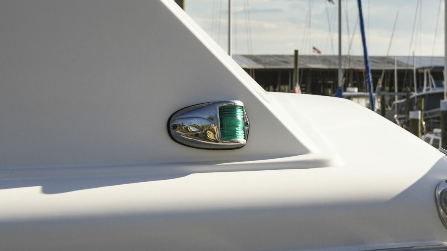 License-free image - Close-up of the side of a white boat featuring a green navigation light encased in a chrome fixture. In the background, there are docks and moored boats, along with a glimpse of the sky and some clouds. The scene suggests a marina setting. A starboard sidelight shown during the daytime aboard a vessel in South Daytona, Florida.